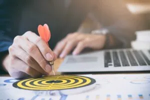 A person in a business suit holding a red dart and placing it on the center of a small yellow and black target, with a laptop and financial documents in the background.